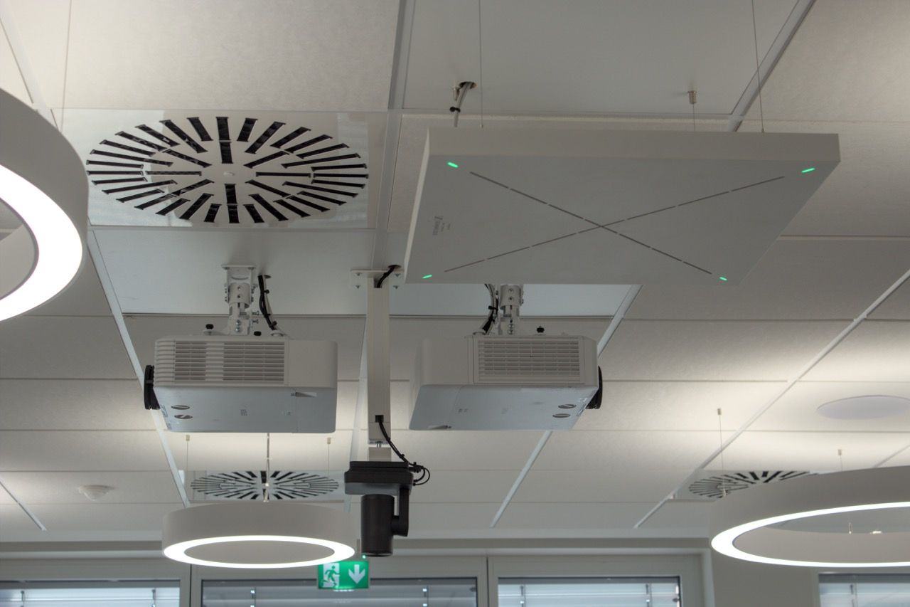 Decke des Konferenzsaals mit zwei Rücken an Rücken montierten Epson-Laserprojektoren, PTZ-Kamera darunter, Sennheiser-Deckenmikrofon und Deckenlautsprecher