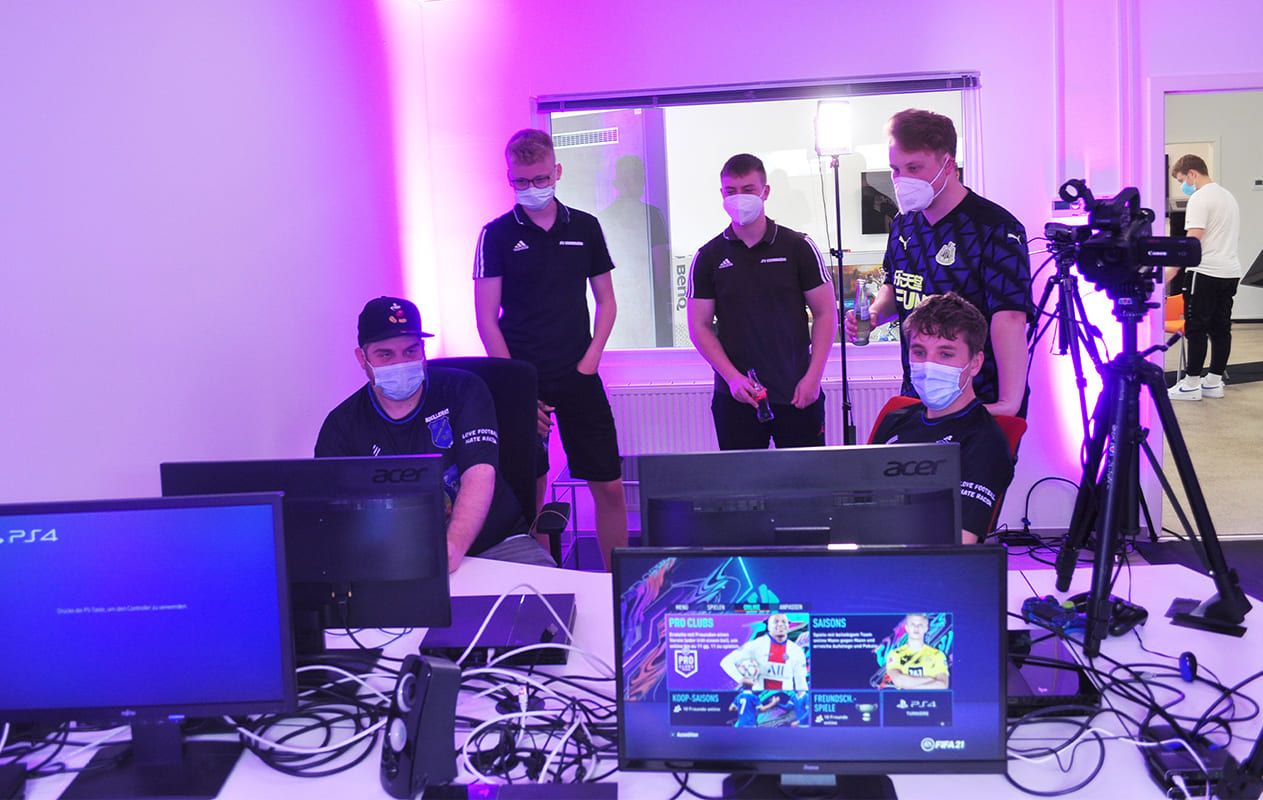 Spieler und Zuschauer an den Spielplätzen mit PS4-Bildschirmen, Kamera auf Stativ und lila Ambientebeleuchtung