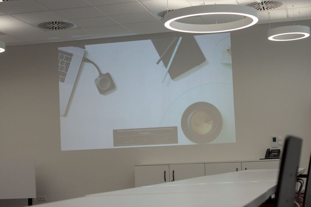 Leinwand mit dem ClickShare-Startbildschirm „Willkommen im Besprechungsraum Feuerwehrhaus Friedeburg“