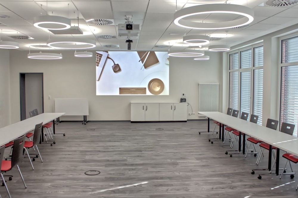 Konferenzsaal im Feuerwehrhaus Friedeburg mit Tischreihen, Projektion an der Stirnwand und runden Pendelleuchten